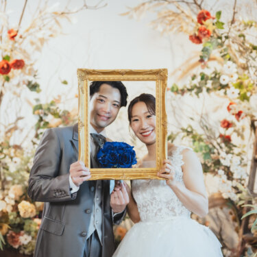 ドライフラワーに囲まれたセットで、金の額縁の中から笑顔を見せる新郎新婦の小物使いのスタジオフォト