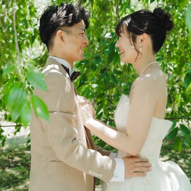 新緑の木の下で抱き寄せ合い、見つめ合って笑う新郎とウェディングドレスの花嫁の結婚写真