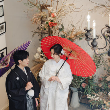 紫と赤の和傘の下で見つめ合う新郎新婦。燭台とドライフラワーがクラシカルな和装スタジオフォト