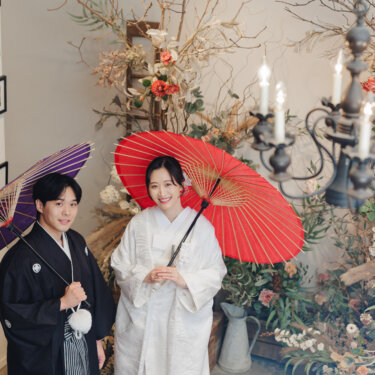 燭台と額縁の並ぶ壁の前で、紫と赤の和傘を差してほほえむ紋付袴の新郎と白無垢の花嫁の和装前撮り