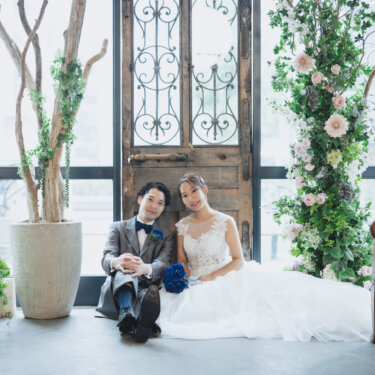アンティークの扉と花のアーチのセットで床に並んで座る新郎新婦。青いブーケのナチュラルなスタジオフォト