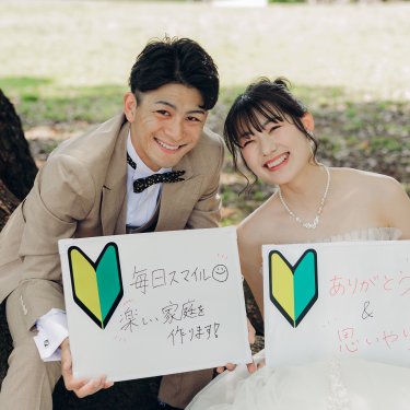 初心者マークと手書きのメッセージボードを持ち、木の根元で笑う新郎新婦。結婚報告にも使えるウェディングフォト