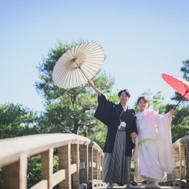 木の反り橋の上で白と赤の和傘を高く掲げる新郎新婦を見上げて写した、白鳥庭園の和装ロケーションフォト