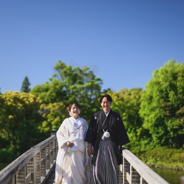 青空の下、池に架かる木の反り橋を並んで歩く白無垢の花嫁と紋付袴の新郎。白鳥庭園の和装前撮り