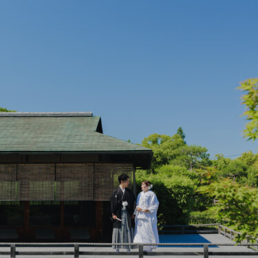 青空の下、池に張り出した数寄屋の建物の縁に立つ白無垢の花嫁と紋付袴の新郎。日本庭園の和装ロケーションフォト