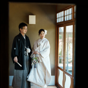 数寄屋の建物の中で、ブーケを手に並んで立つ紋付袴の新郎と白無垢の花嫁を暗がりから写した和装前撮り