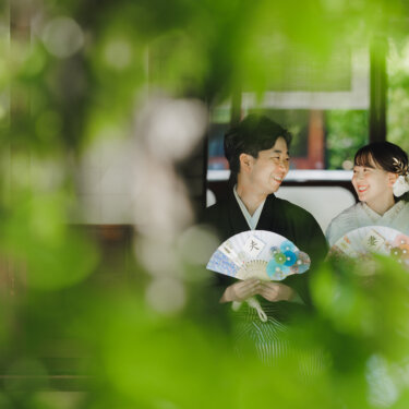 青もみじを前景にぼかし、縁側で'夫''妻'の扇子を持って笑い合う新郎新婦の和装前撮り