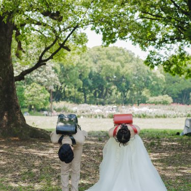 大きな木の下でランドセルを背負い、深くおじぎをするタキシードの新郎とウェディングドレスの花嫁。ユニークな前撮り