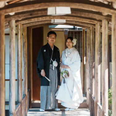 木の柱が続く屋根付きの回廊の奥に並んで立つ、紋付袴の新郎と白無垢の花嫁の和装フォトウェディング