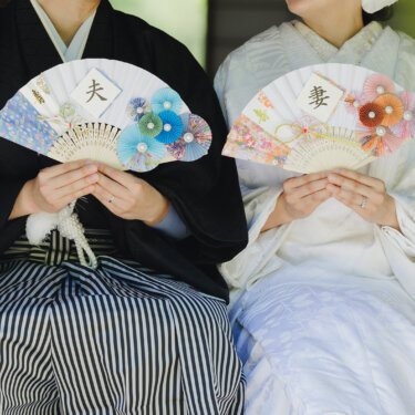 '夫''妻'の扇子を並んで持つ、紋付袴の新郎と白無垢の花嫁の胸元を写したアイテムカットの和装フォトウェディング