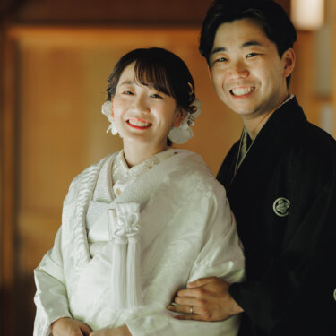 木の廊下で紋付袴の新郎が白無垢の花嫁の腰に手を回し、並んでカメラに笑う和装前撮り