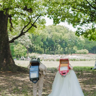 大きな木の下で、ランドセルを背負っておじぎをする新郎新婦の後ろ姿。遊び心ある小物使いのフォトウェディング