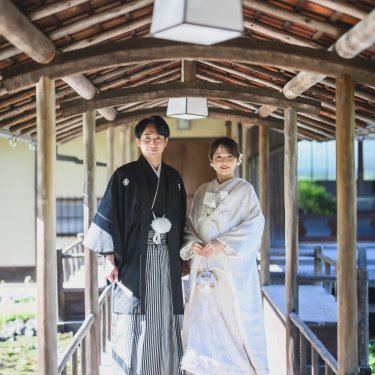 白鳥庭園の屋根付きの回廊に並んで立ち、カメラにほほえむ紋付袴の新郎と白無垢の花嫁の和装前撮り