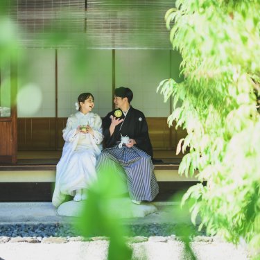 青もみじを前景に、縁側に座って抹茶を楽しむ紋付袴の新郎と白無垢の花嫁の和装前撮り
