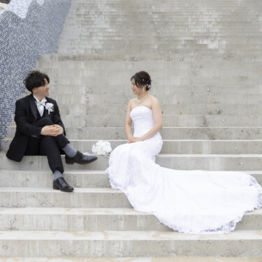 青いタイルの曲線の壁がある海辺の階段に離れて座り、見つめ合う新郎新婦。トレーンが美しいウェディングフォト