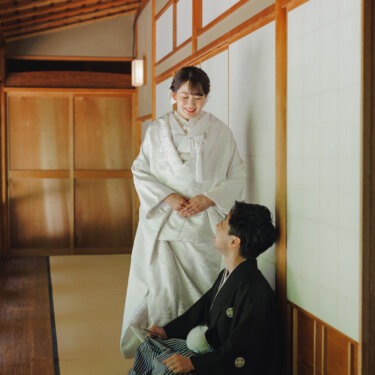 畳の廊下で立つ白無垢の花嫁と、しゃがんで見上げる紋付袴の新郎。日本家屋で撮る和装フォトウェディング