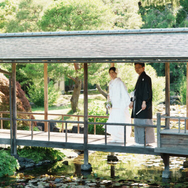 池に架かる屋根付きの廊下に立つ白無垢の花嫁と紋付袴の新郎を、緑の庭園ごと写した和装ロケーションフォト