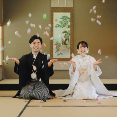 掛け軸のある和室で、折り鶴を舞い上げて笑う紋付袴の新郎と白無垢の花嫁の和装前撮り