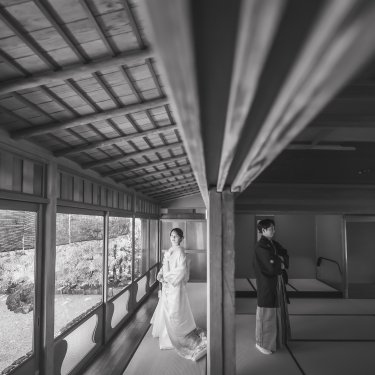 長い縁側と畳の広間に離れて立つ新郎新婦をモノクロで写した、奥行きのある和装前撮り