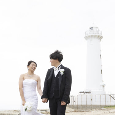 白い灯台の立つ海辺で手をつないで笑い合う、タキシードの新郎とウェディングドレスの花嫁の前撮り