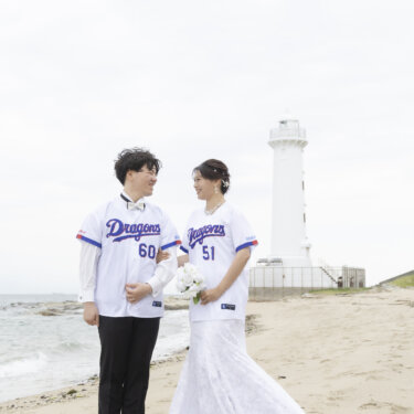 白い灯台を背に、野球のユニフォームを羽織った新郎新婦が砂浜で見つめ合う楽しいロケーションフォト