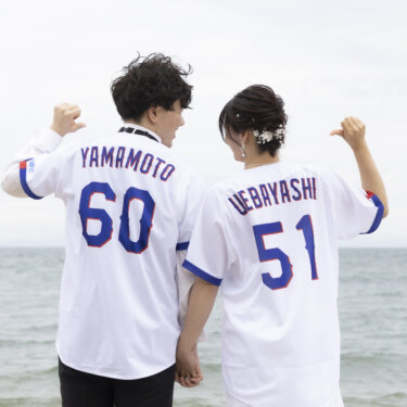 背番号入りの野球のユニフォームを着て、海を背に背中の名前を指さす新郎新婦の後ろ姿。遊び心ある前撮り