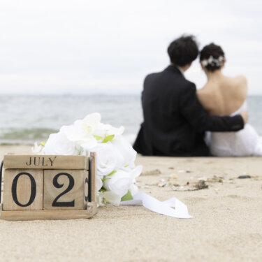 砂浜の日付ブロックにピントを合わせ、奥で肩を寄せる新郎新婦をぼかした海辺のウェディングフォト