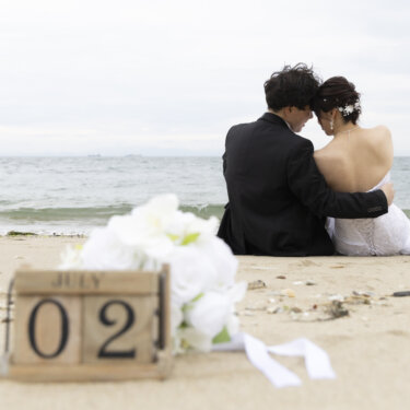 砂浜に置いた日付ブロックと白いブーケの奥で、海を眺めて寄り添う新郎新婦の後ろ姿の前撮り