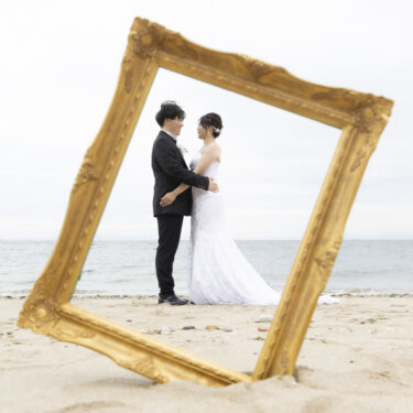 砂浜に立てた金の額縁越しに、海を背に抱き合う新郎新婦を写した遊び心ある構図のウェディングフォト
