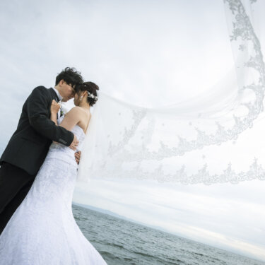 海風にロングベールをなびかせて抱き合い、キスをする新郎新婦。海辺で撮るドラマチックな前撮り