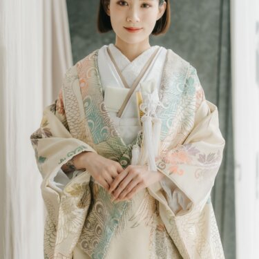 淡いクリーム地に孔雀と花模様を金糸で織り出した色打掛をまとい、手を重ねて微笑む花嫁。スタジオでの和装前撮り