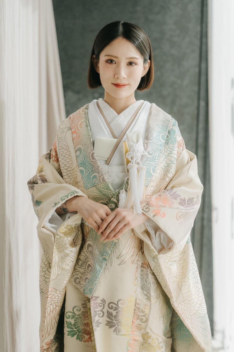 淡いクリーム地に孔雀と花模様を金糸で織り出した色打掛をまとい、手を重ねて微笑む花嫁。スタジオでの和装前撮り