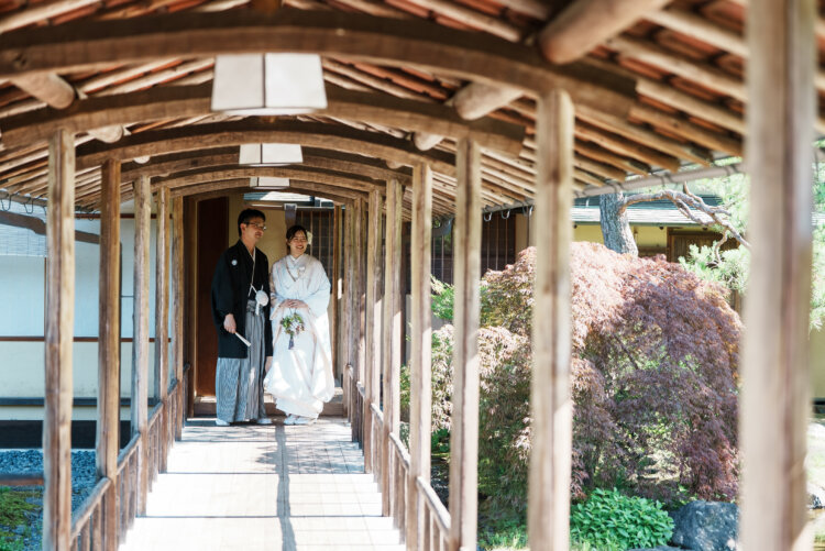 白鳥庭園の屋根付きの回廊に立つ新郎新婦を、紅葉した木とともに写した和装ロケーションフォト
