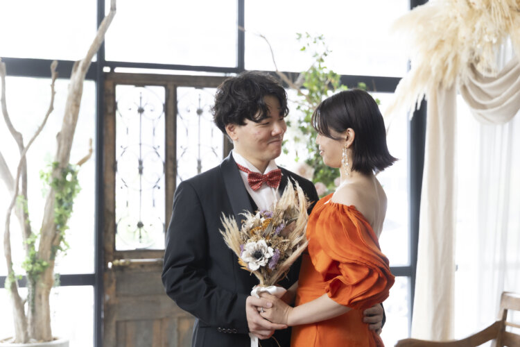 アンティークの扉の前で手を取り合い、見つめ合う新郎とオレンジのカラードレスの花嫁。ドライフラワーのブーケの結婚写真