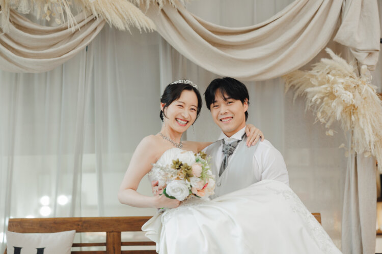 パンパスグラスとドレープのセットで、花嫁をひざに乗せて笑う新郎。ティアラとブーケのウェディングフォト