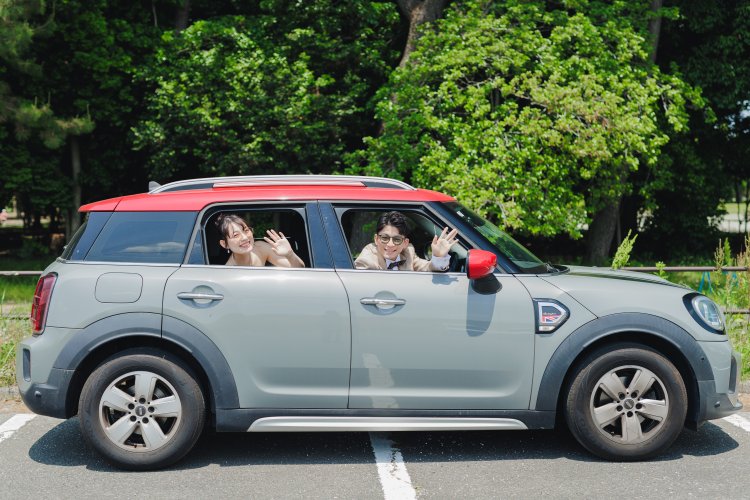 赤いルーフの愛車の窓から手を振るタキシードの新郎とウェディングドレスの花嫁を、車ごと写した遊び心ある前撮り